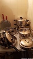 Various stainless steel pots and pans with lids arranged on tabletop, showing utensils and cookware details.