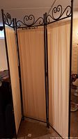Full view of the three-panel room divider showing black ornate metal frame and cream cloth panels in upright position.