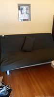 Futon sofa with black mattress cover and toss cushion sitting on hardwood floor with a framed art picture above.