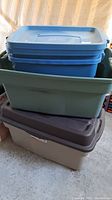 Four plastic tote bins stacked and arranged in garage area. Blue and gray bins on top of larger brown/gray tubs.
