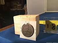 Front view of a metal token or charm with a stylized "OM" monogram mounted in paper holder.