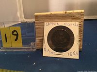 Front view of the 1854 Upper Canada half penny coin placed in a protective holder with handwritten labels and part of a plastic container.