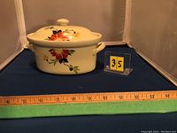 Front view of the small oval Hall USA casserole dish with lid on a dark blue surface next to a measuring tape indicating approximately 10 inches in length.