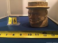Front view of jug showing detailed face, hat lettering, and approximate height using a tape measure and yellow evidence marker '16'.