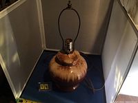 Front view of a vintage pottery lamp with beige and brown glaze, metal socket, and harp for lamp shade, sitting on a blue surface with a ruler and item number display.