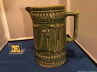 Green pottery jug showing handle and relief decoration on the side.