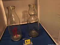 Photo showing two vintage glass milk bottles: on left, a bottle embossed with a red Silverwoods logo; on right, a cream top bottle dated 1923. Both set against a neutral background with yellow lot number 31.