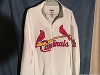 Front view of a white zip-up jacket with large St. Louis Cardinals logo across the chest and embroidered emblem on the sleeve, hanging on a hanger.