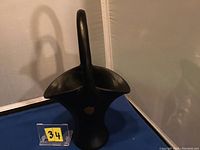 Photo showing the front and slight side view of a black satin glass basket with a tall curved handle, sitting on a dark blue surface with a yellow lot number card beside it.