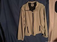 Full view of the khaki jacket hung on a hanger showing front zipper and pockets.