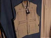 Front view of khaki vest with multiple pockets and metal rings, hanging on a hanger against blue curtain.