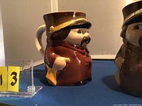 Left side view of a brown and yellow glazed ceramic Goebel stein shaped like a train conductor with mustache and hat, approx. 5 inches tall.