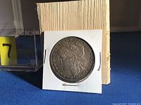 Front side of 1880 Morgan Silver Dollar showing Lady Liberty profile in white cardboard holder against a beige background.