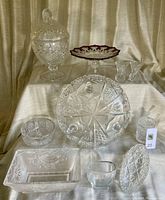 Overview of ten assorted cut glass and crystal pieces on tiered white cloth background against beige drape