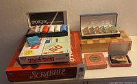 Shows Scrabble unopened box, Monopoly board game with property cards, poker chip set inside box, small box of tokens, and wooden card holders with fabric insets on a wooden stand.
