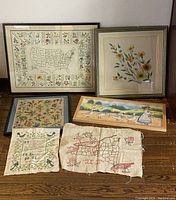 Overview of all six hand embroidered needlework pieces including US states map, floral embroidery, Irish blessing prayer, and girl feeding ducks scene.