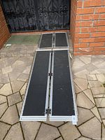 Full view of the two wheelchair ramps unfolded on outdoor paving, showing the fold-in-half hinge and the non-slip black surface.