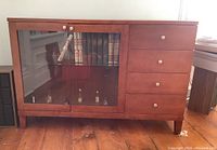 Front view of wood and laminate cabinet with double glass doors and four drawers, showing contents and visible wear and scratches.