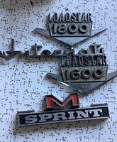 Four metal car badges placed on a speckled background, showing Loadstar 1800, Loadstar 1600, M Sprint and International badges.