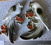 Pair of chalkware decorative birds lying flat against a patterned background, showing full views of both birds, detailing the colors and floral elements held by the birds.