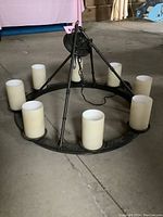 Round metal chandelier frame showing eight frosted cylindrical glass light shades around the ring. It is placed on the floor, showcasing the chain and canopy. The metal has some dust and wear.