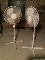 Front view of two white oscillating floor fans side by side on stand bases on a concrete floor.