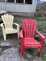 Two plastic Muskoka chairs in red and cream colors placed outdoors on ground with rocks and dry grass.
