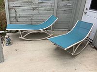 Pair of blue mesh patio rocking loungers with white metal frames on concrete patio outdoor area, showing overall size and shape.
