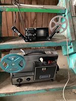Side view of the film projector on the lower shelf with turquoise reel, and film editor on the upper shelf with one clear reel.