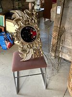 Ornate novelty wall clock with gold-tone molded plastic frame featuring winged figures and roses, black clock face, resting on pink-upholstered chair seat.