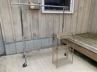 View of both items outdoors near a wooden wall: chrome metal clothing rack and gold-tone metal record holder.