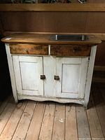 Full front view of the antique wood cabinet showing the white painted lower section with two cabinet doors, the natural wood countertop with metal sink insert on the right, and two drawers on the top with worn wooden knobs.