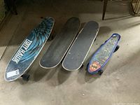 Four skateboards arranged on the floor showing their top sides, including Tony Hawk skateboard with blue feather wing graphic and two standard skateboards with black grip tape.