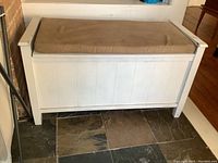 White wood storage bench with hinged lid and light brown micro suede seat pad, placed on a tiled floor.