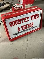 Front and angled view of rectangular plastic store sign reading 'COUNTRY TOYS & THINGS' with a red plastic border showing wear and scratches.