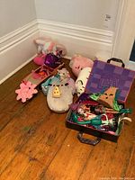 Photo of assorted plush toys, toy car, Monster High dolls, and carry case, placed on wooden floor against white wall