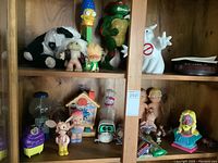 Shelf with various toys showing the giant Marge Simpson Pez dispenser, black and white plush cow, Troll dolls, a ghost figure, and small figures.