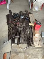 Overview of several metal model train tracks laid on floor with a wooden board holding a red toy transformer and electrical controller. Wires scattered among tracks.