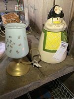 Image of the cookie jar and lamp placed side by side on a rustic wooden surface, showing details of the lamp's floral glass shade and the Babooshka cookie jar figure.