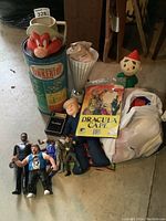 Wide photo of lot items spread on floor, showing Yosemite Sam mug, Tinkertoy container, Cracker Jack doll, Beano comic book, various action figures.