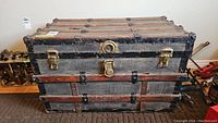 Front view of antique steamer trunk showing brass latches, central lock, wooden slats, and leather pulls with one missing pull.