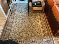 Wool area rug laid out on floor showing full rug with visible wear, stains, and discoloration near couch and box.