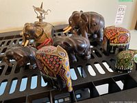 Collection of seven elephant figurines of varied material and finishes on a black metal surface.