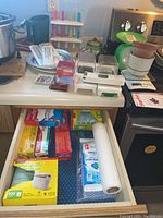 Counter with Starfrit kitchen scale, alligator dicers, popsicle makers in pastel colors, thermometers, plastic green milk jug, and adjacent open drawer filled with various food wrap boxes and compostable bags.