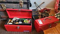 Photo showing three metal tool boxes, one black and two red, with one red Mastercraft tool box open revealing hand tools, hardware items, and bottles inside. Various automotive tools and stands visible behind.