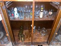 Wooden cabinet with three shelves holding various drinking glasses including clear glasses, amber pedestal glasses, and a white ceramic bottle.