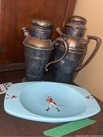 Silver plate cocktail shakers/pitcher decanters with tarnish showing next to a blue Wade dish with sailor figure design on a wooden surface.