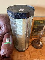 Octagonal glass and plastic Zippo display cabinet next to a leather chair and floor lamp, showing top and side view with visible Zippo logo and clear internal shelves.