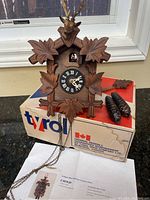 Antique West German wooden cuckoo clock with carved leaves and deer head, resting on original Tyrol box with two pinecone-shaped weights visible. Clock face with Roman numerals and chains hanging down.