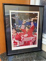 Framed painting showing Canadian hockey players celebrating a championship win with trophy raised, predominantly red and white uniforms.
